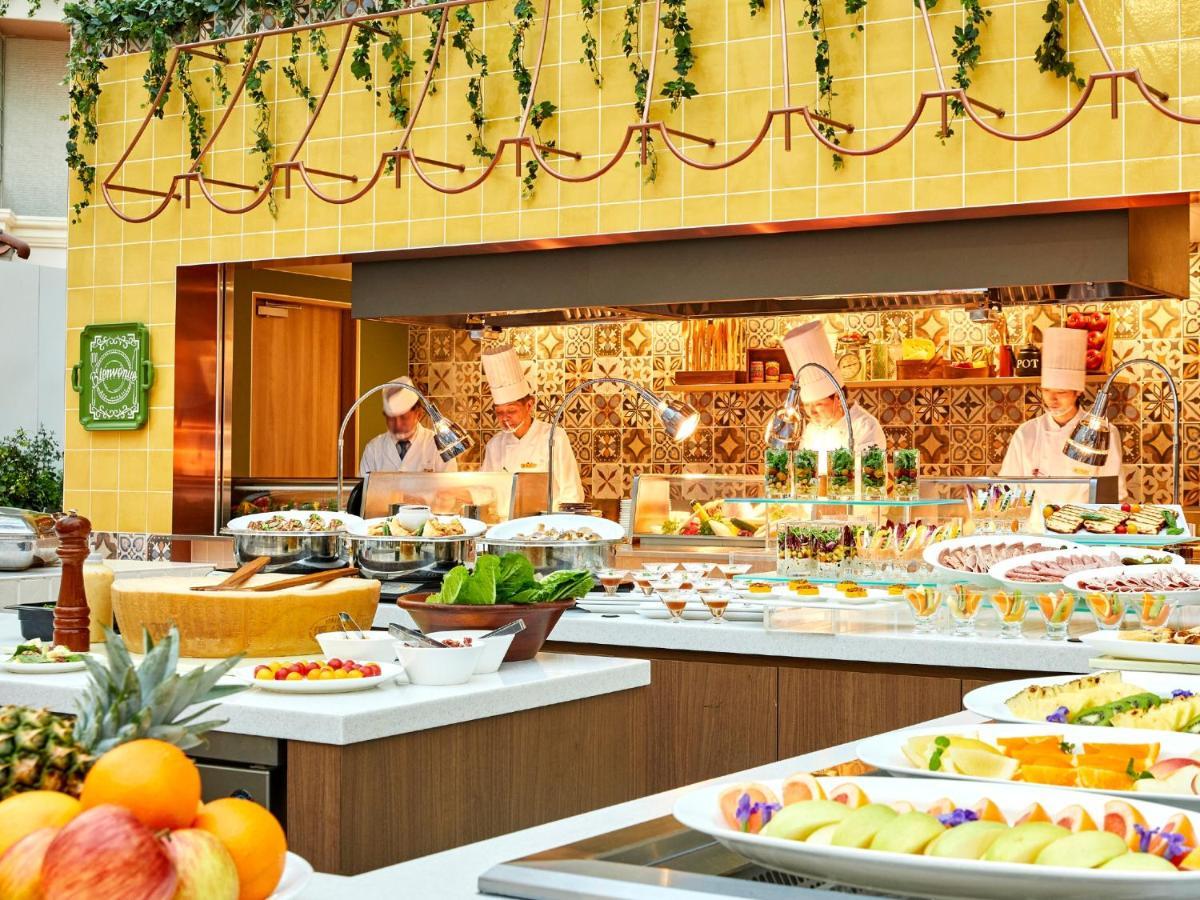 Grand Nikko Tokyo Bay Maihama Otel Urayasu Dış mekan fotoğraf The photo shows a vibrant and colorful buffet setup in a restaurant. In the foreground, there are various dishes arranged on white platters, featuring an assortment of fruits, desserts, and other items. The background features chefs in white uniforms