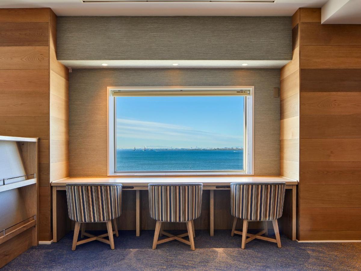 Grand Nikko Tokyo Bay Maihama Otel Urayasu Dış mekan fotoğraf The photo shows a modern workspace with three striped chairs positioned at a long, white desk. The desk faces a large window with a view of a tranquil body of water and a distant skyline. The walls around the workspace are made of wood paneling, crea