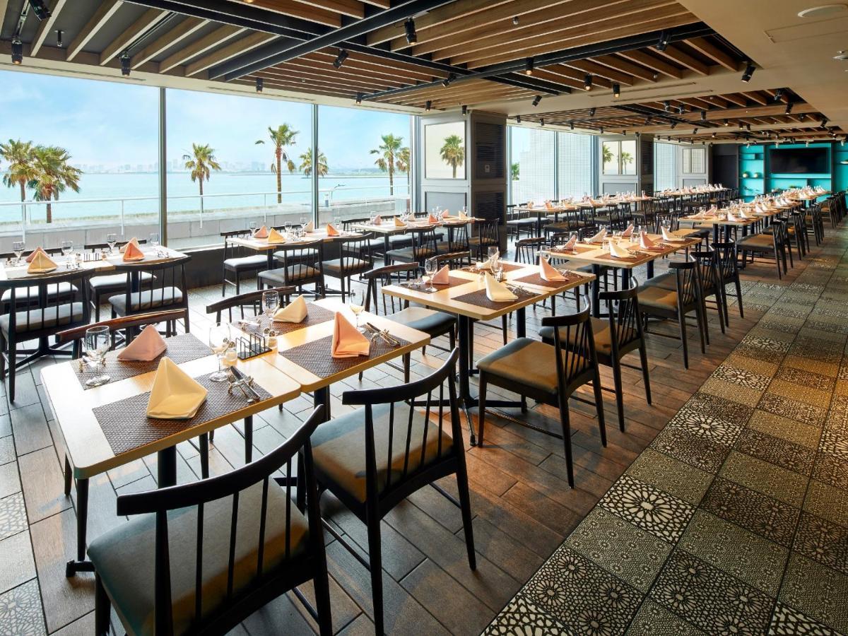 Grand Nikko Tokyo Bay Maihama Otel Urayasu Dış mekan fotoğraf The photo shows a modern restaurant interior featuring rows of neatly arranged tables and chairs. The large windows provide a scenic view of palm trees and what appears to be a body of water in the background, suggesting a coastal location. Each tabl