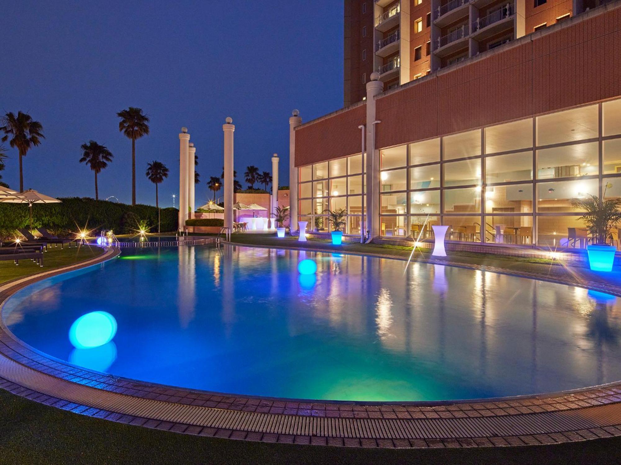 Grand Nikko Tokyo Bay Maihama Otel Urayasu Dış mekan fotoğraf The photo shows a well-lit swimming pool at night, surrounded by a lush green area. The pool features glowing blue lights that create a serene and inviting atmosphere. Palm trees can be seen in the background, adding a tropical feel to the scene. Add