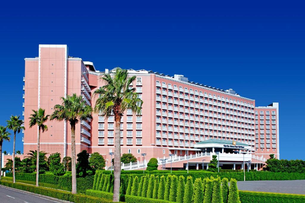 Grand Nikko Tokyo Bay Maihama Otel Urayasu Dış mekan fotoğraf The image shows a large, multi-story hotel building featuring a light pink facade. The architecture appears modern, with numerous windows arranged in a grid pattern. In the foreground, there are neatly trimmed shrubs and palm trees, adding a tropical