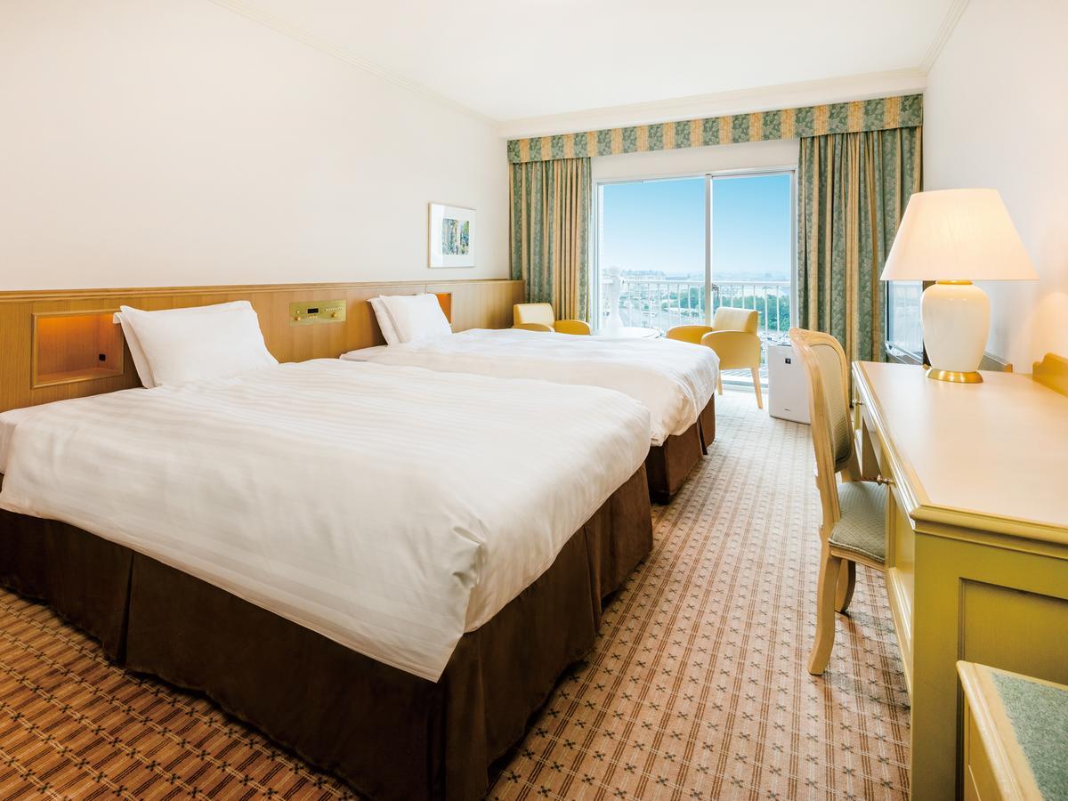 Grand Nikko Tokyo Bay Maihama Otel Urayasu Dış mekan fotoğraf The photo shows a hotel room featuring two twin beds with white linens and dark brown bed skirts. The room has a light, neutral-colored wall and carpet. There is a window with curtains that let in natural light, offering a view outside. A small desk 
